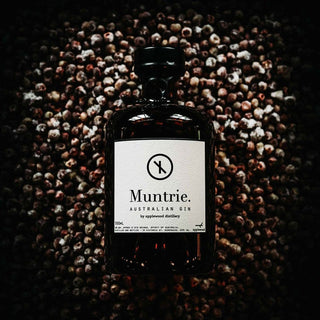 A dark glass bottle of Applewood Muntries Gin 500ml with a white label rests on a surface scattered with small round brown muntries berries, dramatic lighting highlighting its infusion of native Australian botanicals.
