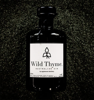 A dark glass bottle of Applewood Davidson Plum Gin 500ml rests on a textured green surface. Its minimalist label has black text, an abstract leaf logo, and highlights native botanicals such as juniper and Davidson plum.
