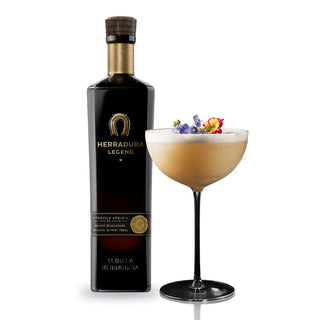 A bottle of Herradura Legend Anejo Tequila 750ml stands beside a coupe glass with a light brown cocktail, topped with colorful edible flowers. Vanilla and toasted oak notes evoke the tequila’s rich character against a white background.