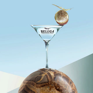 A martini glass with "Beluga" labeling sits on polished brown marble, adorned with a gold garnish, set against a light blue background—evoking the floral malt notes and smooth finish of Beluga Celebration Limited Edition Vodka 700ml.