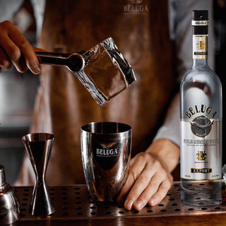 A bartender drops a large ice cube into a metal shaker. Next to it sits a 1.5L bottle of Beluga Noble Russian Vodka on the bar, known for its creamy smoothness and grain sweetness, among various bartending tools.