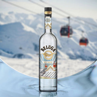 A bottle of Beluga Noble Winter Limited Edition Vodka 700ml stands upright on a reflective icy surface, with snowy mountains and two red cable cars in the blurred background, emphasizing its smooth palate and refined character.