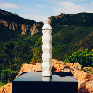 White geometric sculpture in a natural landscape with mountains and greenery.