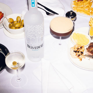 A table set with Belvedere Vodka & Bar Spoon 700ml, a martini glass, olives, fries, steak with sauce, and fresh bread on a white tablecloth offers a smooth palate and refined finish to your dining experience.