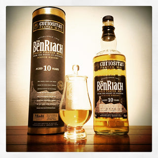 A bottle of Cutiositas Ben Riach aged 10 years single malt Scotch whisky, shown next to its matching cylindrical tin packaging and a small tulip glass filled with the golden whisky, set on a wooden surface against a plain background.