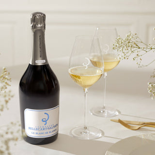 The Billecart-Salmon Le Blanc de Blancs Grand Cru NV + 2 Glasses Gift Set sits on a white table with delicate flowers and gold cutlery, evoking the cuvée’s chalky minerality and citrus elegance in a refined, softly lit ambiance.