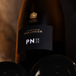 A close-up of the Bollinger PN TX20 Champagne bottle, with its golden label against a dark background, framed by other bottles, emphasizing ripe orchard fruit flavors and refined chalky minerality.