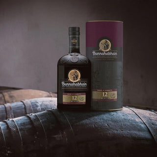 A bottle of Bunnahabhain 12 Year Old Cask Strength Single Malt Scotch Whisky sits by its box on whisky barrels, set against a rustic backdrop—notes of cracked black pepper and creamy vanilla hint at its complex character.