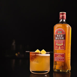 A bottle of Bushmills Red Bush Irish Whiskey sits beside a cocktail garnished with pineapple chunks and spiced rim, set against a dark backdrop, highlighting the whiskey’s caramel and toasted oak notes.