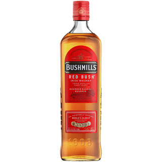 A bottle of Bushmills Red Bush Irish Whiskey with a red label and gold text, displaying the Bushmills logo and “World’s Oldest” claim, stands upright against a white background, suggesting hints of vanilla and toasted oak.