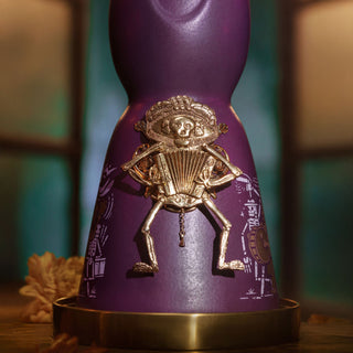 A close-up of Clase Azul Dia de Los Muertos 2024 Música Limited Edition Tequila 1L shows a purple bottle with a metallic skeleton playing an accordion, warm lighting, graphic designs, and notes of cooked agave and toasted oak.