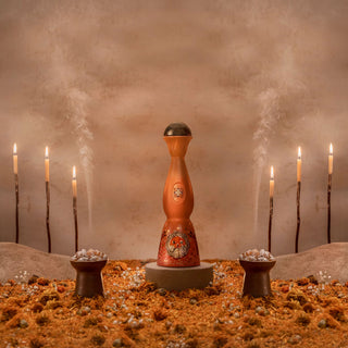 The Clase Azul Dia de Los Muertos 2023 Aromas Limited Edition, an ornate orange incense holder with a tiger design, stands on a pedestal amid two bowls, six candles, vanilla caramel notes, and dried flowers in a neutral setting.