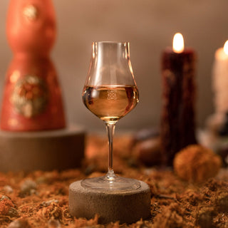 The Clase Azul Dia de Los Muertos 2023 Aromas Limited Edition stands on a round stone base with amber liquid, surrounded by dried foliage and two glowing candles. Vanilla caramel aromas linger, blending into the warm, cozy background.