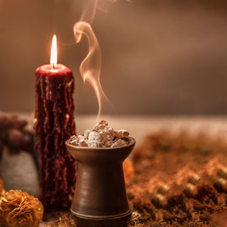 The Clase Azul Dia de Los Muertos 2023 Aromas Limited Edition sits beside a lit red candle with melted wax, a brown incense burner releasing fragrant smoke, dried flowers, and hints of vanilla caramel, set against a warm, blurred background.