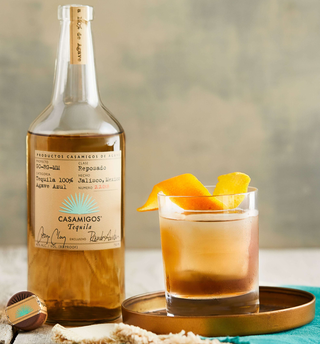 A bottle of Casamigos Reposado Tequila 700ml, celebrated for sweet agave and caramel notes, sits next to a cocktail with orange peel on a bronze tray, alongside its wooden cap and a turquoise napkin.