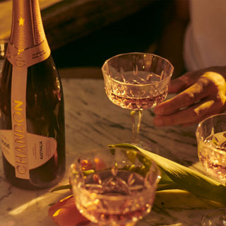 A bottle of Chandon Brut Rosé on a marble table with three filled crystal glasses, a hand holding one, and a tulip beside them in warm light—highlighting its fine bubbles and balanced acidity.