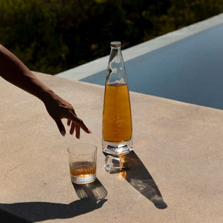 A hand reaches for a glass of amber Cincoro Anejo Tequila 750ml, rich with toasted oak notes, beside its tall, elegant bottle on a sunlit poolside surface, with greenery and shimmering water softly blurred in the background.