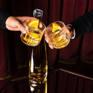 Two hands toast with glasses of golden cocktails and orange slices above a Cincoro Reposado Tequila 750ml bottle, set before a dark red curtain—capturing the spirit’s toasted oak and honey notes in every sip.