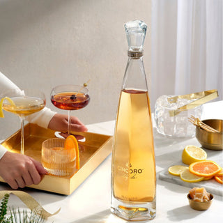 A 750ml bottle of Cincoro Reposado Tequila sits on a white table beside two cocktails, sliced lemons, and a gold bowl; a hand reaches for a drink garnished with orange and notes of caramelized agave.