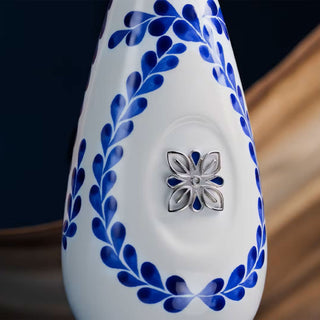 A close-up of the Clase Azul Reposado Tequila 750ml bottle, a white ceramic vessel adorned with a blue leaf pattern and a silver floral ornament, set against a blurred caramel-toned brown background.