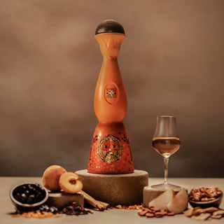 A tall, ornate orange and red Clase Azul Dia de Los Muertos 2023 Aromas Limited Edition bottle sits on a stone pedestal beside a wine glass, surrounded by peaches, berries, nuts, and spices—evoking cooked agave and toasted oak notes.