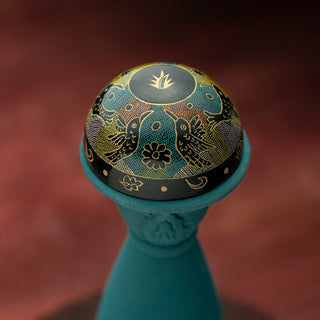 A close-up of Clase Azul Mezcal Guerrero’s decorative round lid, adorned with gold bird and floral patterns on black atop a teal vessel, set against a reddish blur, evokes the honeyed sweetness and allure of roasted agave.