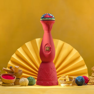 The Clase Azul Mezcal San Luis Potosi 750ml features a tall, decorative red bottle with intricate patterns and a vibrant cap, set before a yellow fan backdrop among bowls of colorful powders, wooden tools, and yarn balls.