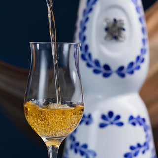 Golden tequila with notes of vanilla is poured into a curved glass. In the blurred background, a Clase Azul Reposado Tequila 750ml bottle—white ceramic with blue floral patterns—adds elegance.