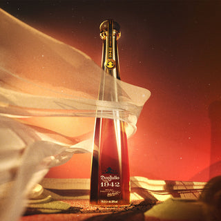 A tall, elegant Don Julio 1942 Añejo Tequila 750ml bottle stands upright against a warm glow, draped in sheer fabric that creates a dramatic, luxurious effect and hints at notes of vanilla and honeyed agave.