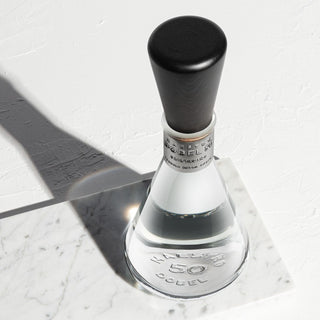A clear, uniquely shaped Maestro Dobel 50 Cristalino Extra Anejo Tequila 750ml bottle with a black cap sits on white marble, displaying "50" and "Dobel," a silver neck band, and notes of sweet agave and toasted oak.