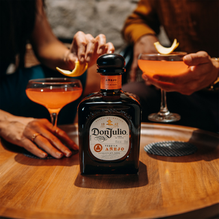 A bottle of Don Julio Anejo Tequila 700ml rests on a wooden table, its agave aroma mingling with orange cocktails topped with orange peel; hands appear in the background as hints of caramel and vanilla fill the air.