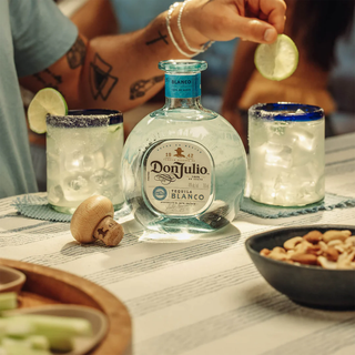 A Don Julio Blanco Tequila 700ml & Shaker Gift Set rests on a striped tablecloth with two glasses of ice and salted rims, a small bowl of nuts, and a hand squeezing lime over a glass for fresh citrus flavor.
