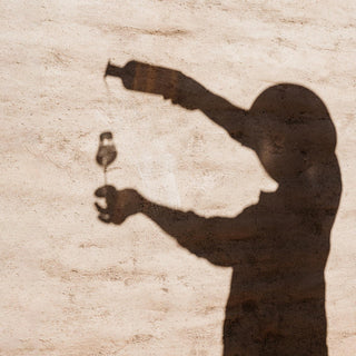 A silhouette pours Don Julio Blanco Tequila 700ml into a wine glass against a textured beige wall, arms raised with bottle and glass, hinting at citrus zest—featured in the Don Julio Blanco Tequila 700ml & Shaker Gift Set.