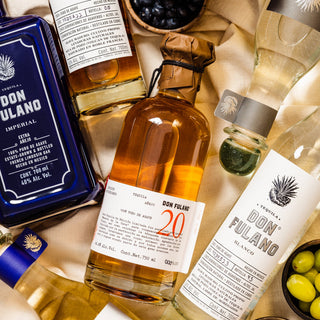A bottle of Don Fulano Imperial Extra-Añejo Tequila 700ml, known for notes of caramel and rich agave, is displayed on a beige cloth beside a bowl of blueberries and a glass of clear liquid.