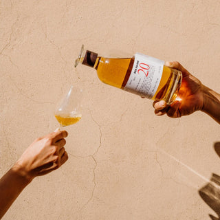 A hand pours Don Fulano 20th Aniversario Sherry Cask Añejo Tequila 700ml from a bottle labeled “20” into a glass held by another hand, with bright sunlight highlighting the scene against a textured beige wall.
