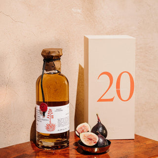 A bottle of Don Fulano 20th Aniversario Sherry Cask Anejo Tequila 700ml, sealed in wax, stands beside a beige box marked with “20”; fresh figs rest nearby on a wooden surface, evoking roasted agave against a textured beige wall.