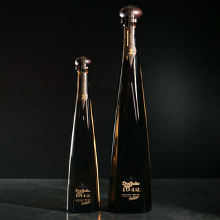 Two tall bottles of Don Julio 1942 Añejo Tequila 1.75L, with notes of roasted agave and oak, stand side by side on a dark surface against a black background. Their amber glass features gold labels and elegant, tapered necks.
