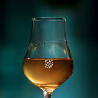 A close-up of Clase Azul Dia de Los Muertos 2024 Música Limited Edition Tequila 1L showcases its amber hue in a wine glass with white floral design, set against a blurry teal backdrop. Notes of toasted oak and vanilla add warmth and richness to the scene.
