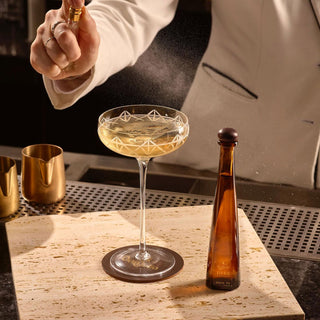 A bartender in a white jacket sprays a fine mist over a champagne coupe with a golden drink; the Don Julio 1942 Añejo Tequila 50ml, in its tall amber bottle, stands nearby on a light marble surface.
