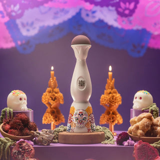 A tall, elegant Clase Azul Dia de Los Muertos 2022 Colores Limited Edition Tequila 1L bottle with a painted skull stands on an altar of candles, sugar skulls, marigolds, traditional breads, fresh agave, and colorful papel picado.