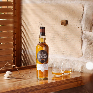 A bottle of Glengoyne 10 Year Old Single Malt Scotch Whisky sits on a wooden table with two partially filled glasses, set against a backdrop of a stone wall and wooden blinds.