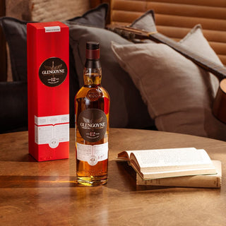 A bottle of Glengoyne 12 Year Old Single Malt Scotch Whisky stands by its red box on a wooden table next to an open book, with sofas and a guitar visible in the background.