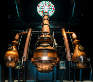Large copper stills in a distillery setting with stained glass window above.