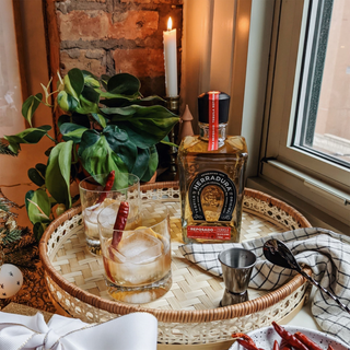 A bottle of Herradura Reposado Tequila 700ml rests on a woven tray with two ice-filled glasses, chili peppers, a metal jigger, napkin, greenery, and a lit candle against a brick wall backdrop.