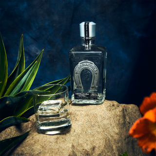 A bottle of Herradura Ultra Añejo Cristalino Tequila 750ml stands on a rock next to a glass of tequila with ice, surrounded by green leaves and an orange flower. Sweet agave and caramel aromas linger against the dark blue background.