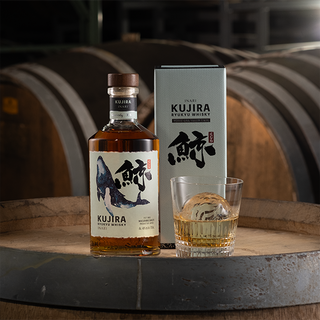 A bottle of Kujira Ryukyu Inari Japanese Whisky, its box, and a glass with whisky and an ice sphere sit on a wooden barrel, with tropical fruit and oak notes evoking the large aging barrels blurred in the background.
