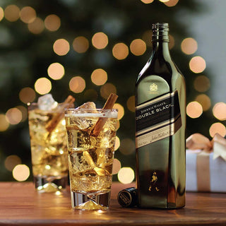 Johnnie Walker Double Black bottle with a glass of whiskey and cinnamon sticks on a wooden table with blurred lights in the background.