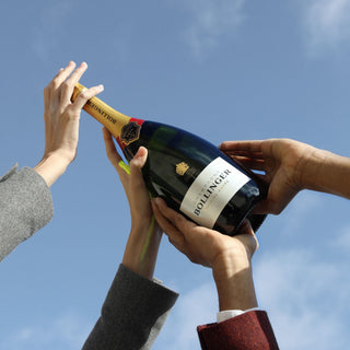 Four hands raise a bottle of Bollinger Special Cuvée Brut NV 750ml (6 Bottles) against a clear blue sky, capturing a festive and joyful moment.