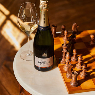 A bottle of Henriot Brut Souverain NV and a filled wine glass rest on a round white table beside a wooden chessboard with arranged pieces, all bathed in warm sunlight that highlights the sparkling wine’s fine bubbles.