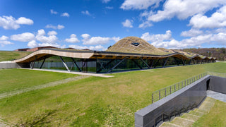 Modern building with a unique roof design on a grassy field under a blue sky with clouds.